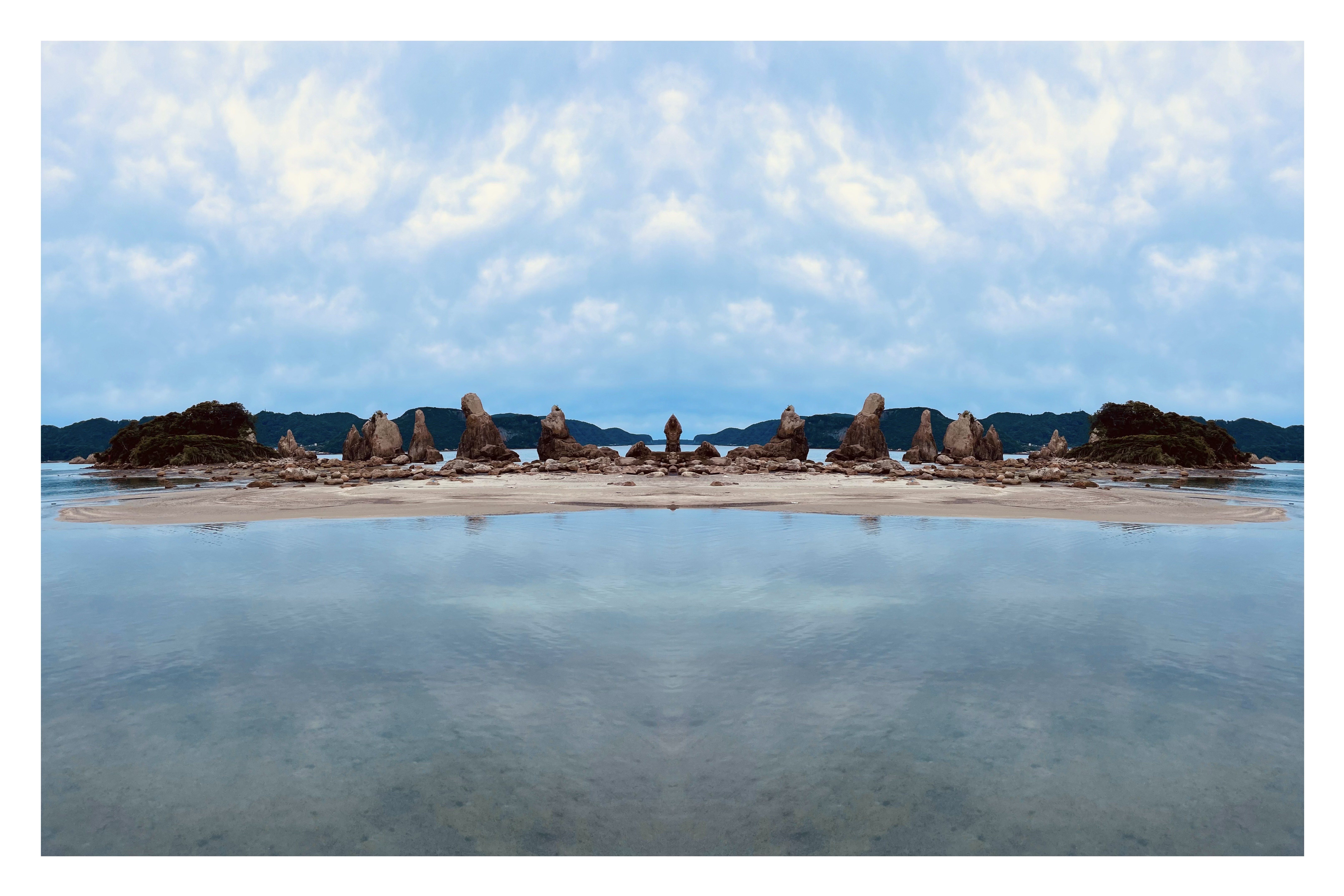 透明度の高い海面が空を映し出す岩場の海岸を左右反転し、奇岩が対称に連なる静謐な構図を生み出した反転写真作品